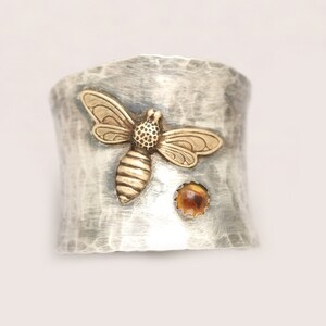 Puede incluir: Un anillo de plata con textura martillada presenta una abeja de oro y una peque&ntilde;a piedra &aacute;mbar.