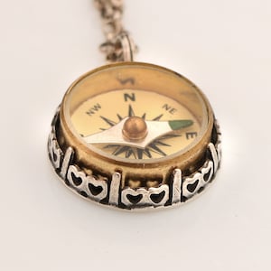 May include: A gold compass pendant necklace with a silver chain and a heart-shaped border. The compass face is white with black markings and a green arrow.