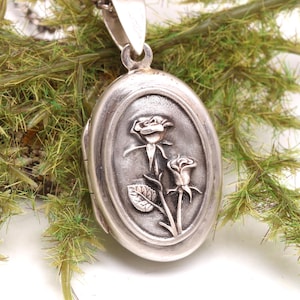 May include: An oval-shaped silver locket pendant featuring a raised relief design of two roses on the front surface. The locket is attached to a silver chain and has a polished metallic finish with darkened recessed areas to highlight the floral detail.