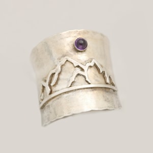 May include: A silver ring with a mountain range design and a small purple amethyst stone set in the center.