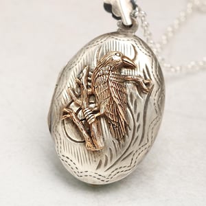 May include: Silver locket necklace with an oval-shaped pendant featuring an intricate design and a gold-toned raven perched on a branch.