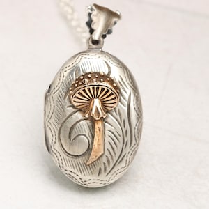 May include: Silver locket necklace with an oval pendant featuring an engraved floral design and a gold mushroom charm.
