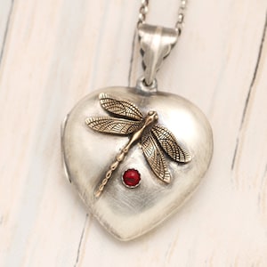 May include: A silver heart-shaped locket with a gold dragonfly design and a red gemstone accent. The locket is on a silver chain.
