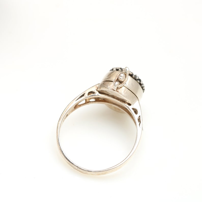 May include: A silver ring with a round, hinged compartment on top. The compartment is decorated with black stones.
