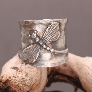 Puede incluir: Un anillo de plata con un diseño de libélula. El anillo es ancho y tiene una textura martillada.