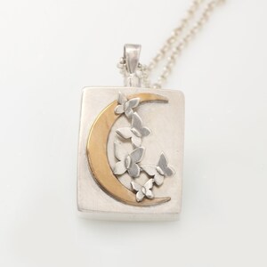 Op de afbeelding: Een zilveren ketting met een vierkante hanger met een halve maan met een gouden afwerking en een ontwerp van zilveren vlinders eromheen.