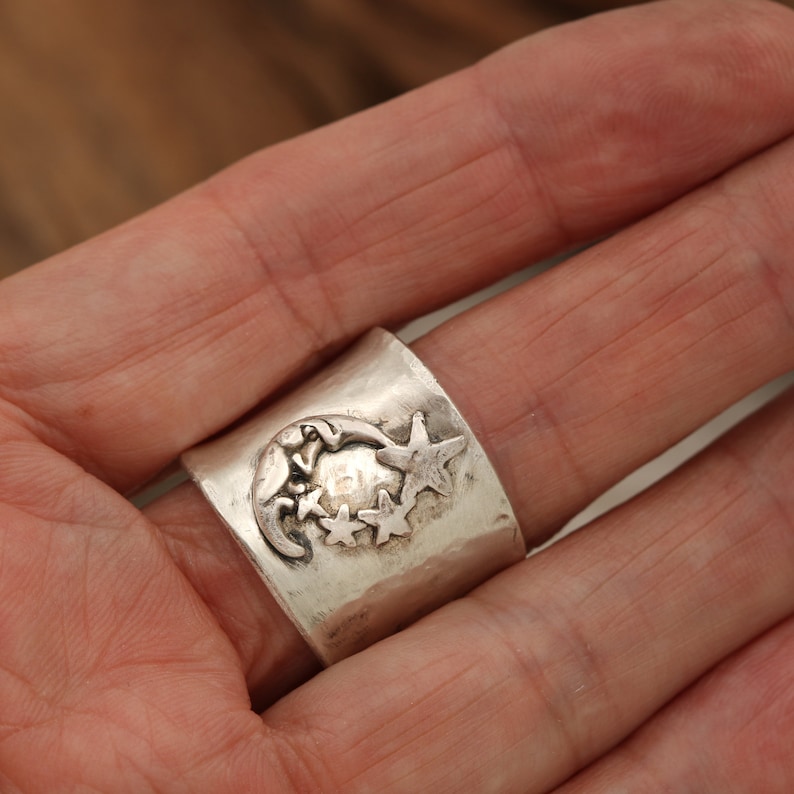 May include: A silver ring featuring a crescent moon and three stars design. The ring is worn on a finger.