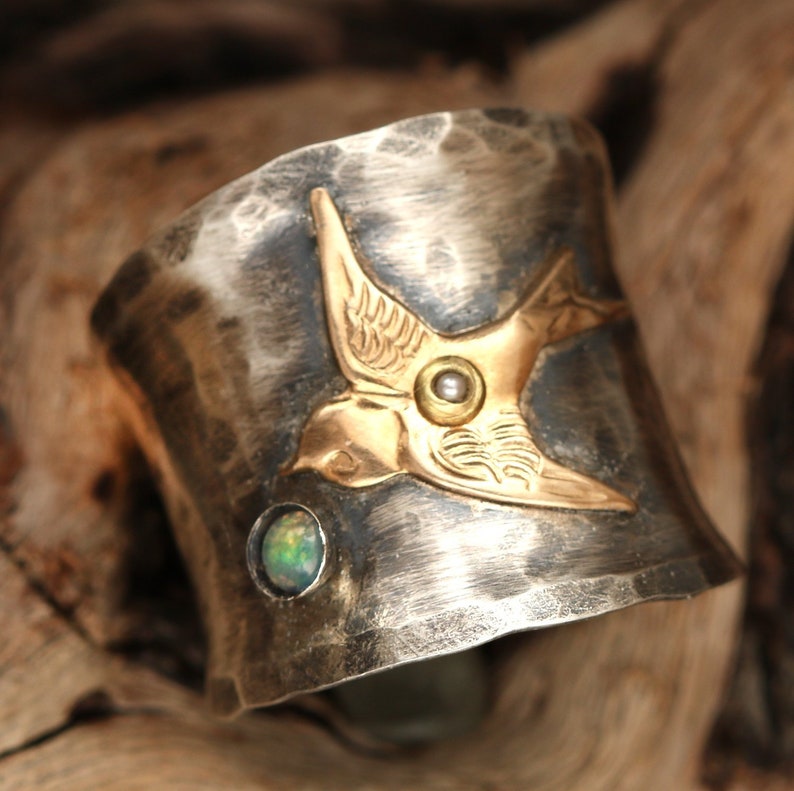 May include: A silver cuff bracelet with a hammered finish features a gold-toned bird design with an opal gemstone. The bird has a pearl-like accent in the center of its chest.
