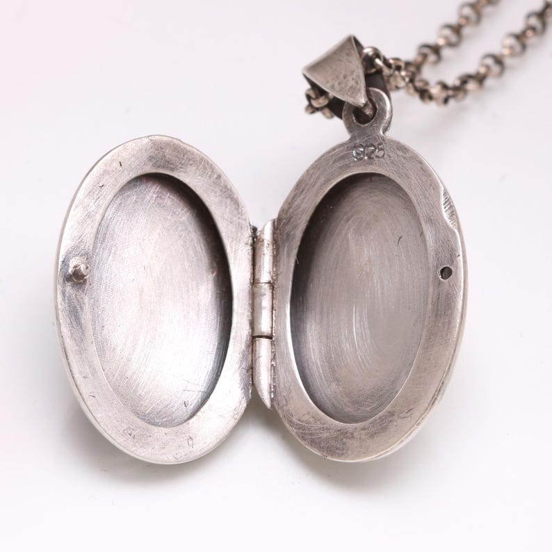 May include: An oval-shaped silver locket pendant with a hinged opening, revealing two empty interior compartments. The metal surface shows a brushed texture and the stamp 925 is visible near the top hinge. A silver chain is attached to the bail.