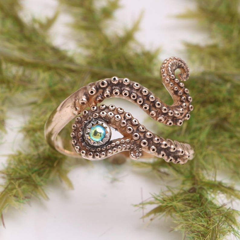 May include: A copper-toned metal ring shaped like a coiled octopus tentacle with textured suction cups along the surface. The center of the spiral features a small, round, iridescent gemstone that reflects light with blue and green hues.