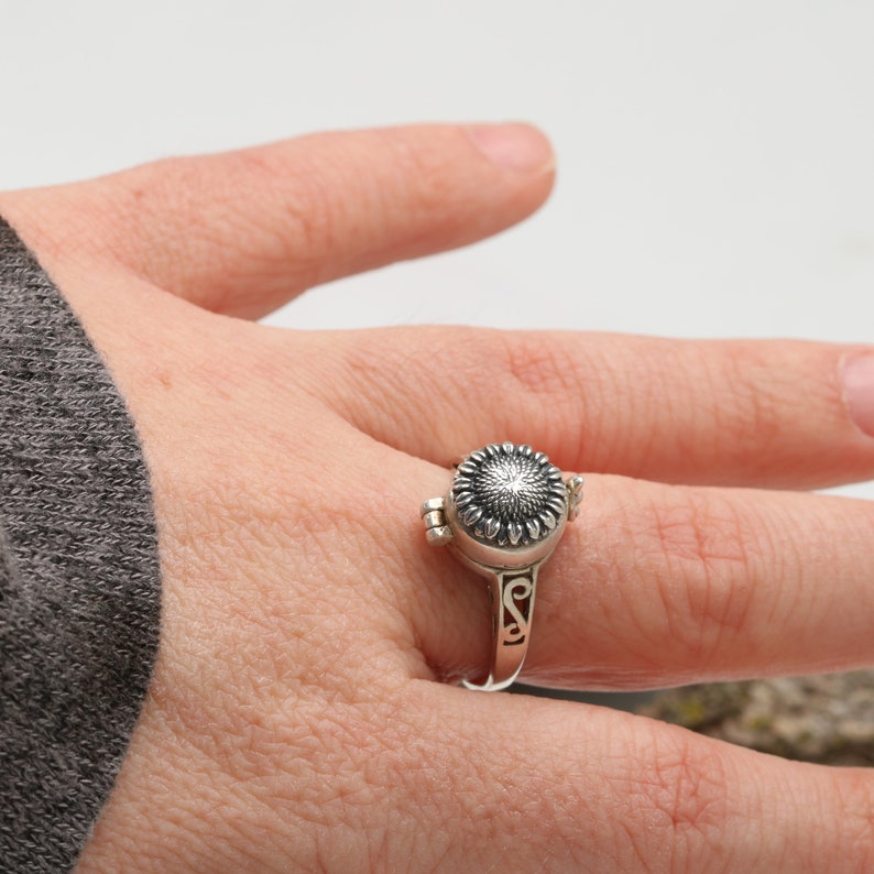 May include: A silver ring featuring a sunflower design on the top. The ring is openable and can be used to hold a small item, such as a photo or a lock of hair.