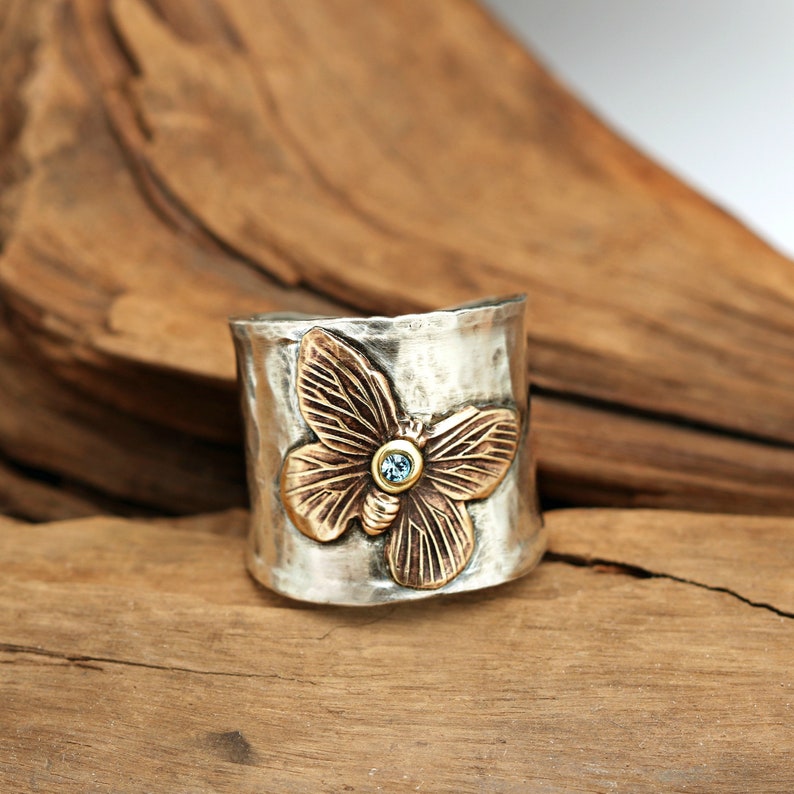 A sterling silver wide band about 3/4 inch wide on top and tapers to 1/2 inch wide underneath the finger.  On the top is a brass butterfly that is angled and soldered onto the ring.  In the center of the butterfly is a small blue Swarovski crystal.