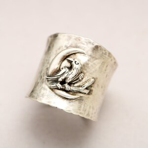 K&ouml;nnte beinhalten: Ein silberner Ring mit geh&auml;mmerter Oberfl&auml;che zeigt ein detailliertes Design eines Vogels, der auf einem Ast in einem Halbmond sitzt.