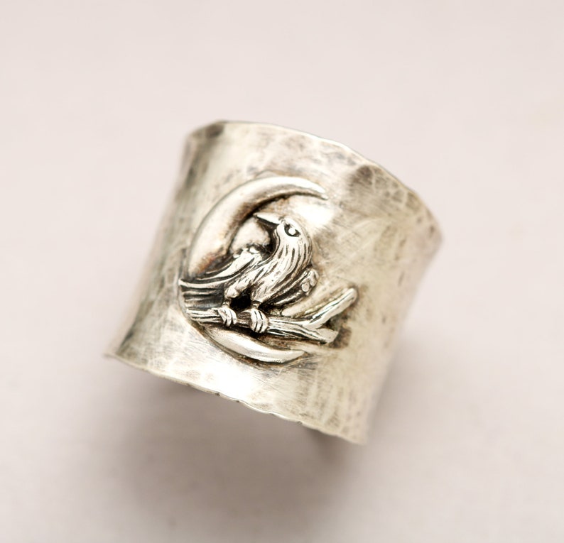 K&ouml;nnte beinhalten: Ein silberner Ring mit geh&auml;mmerter Oberfl&auml;che zeigt ein detailliertes Design eines Vogels, der auf einem Ast in einem Halbmond sitzt.