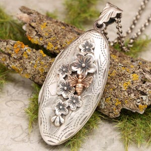 May include: Silver locket necklace with an oval pendant featuring a bee and floral design. The bee is made of gold and the flowers are silver.