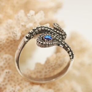 May include: A silver ring shaped like an octopus tentacle with a textured surface featuring small circular suckers. The tentacle wraps around to display a single, small, round, iridescent blue gemstone set securely at the end of the band.