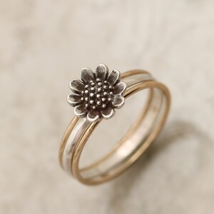 May include: A silver ring with a sunflower design, set on a gold band. The ring is made of two bands, one silver and one gold, that are intertwined.