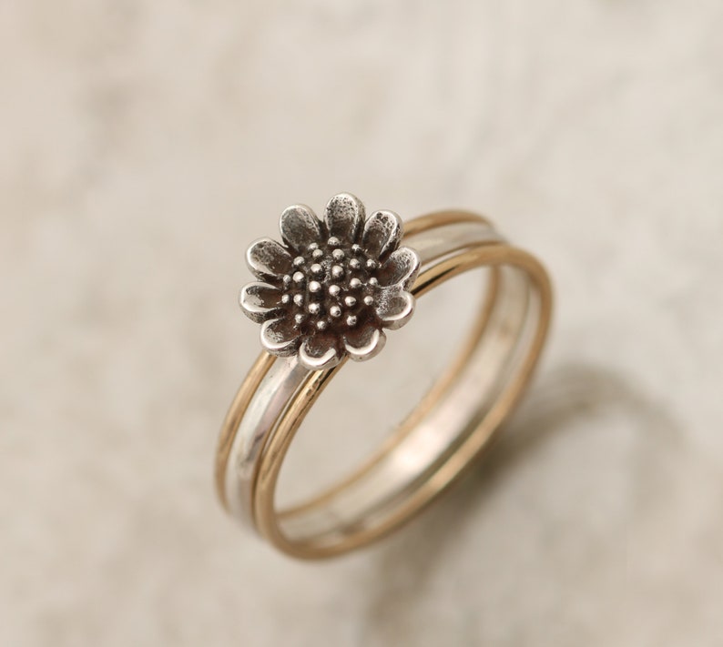 May include: A silver ring with a sunflower design, set on a gold band. The ring is made of two bands, one silver and one gold, that are intertwined.