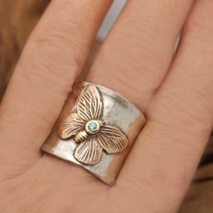 K&ouml;nnte beinhalten: Ein Silber- und Goldring mit einem Schmetterlingsdesign. Der Schmetterling hat einen blauen Edelstein in der Mitte.