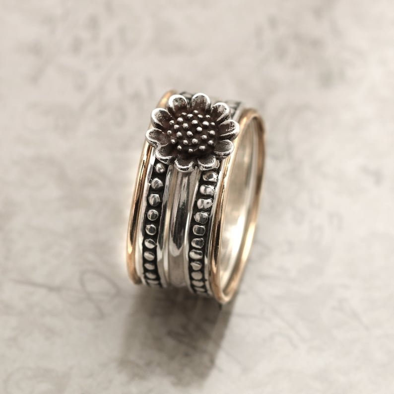 May include: A silver and gold ring with a sunflower design. The ring is made of three bands, each with a different texture. The top band features a sunflower with a brown centre and silver petals. The middle band is smooth and silver. The bottom band is textured with small silver beads.