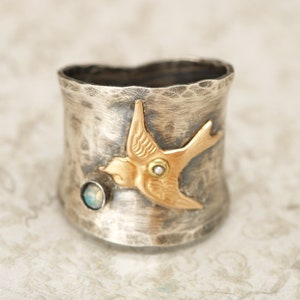May include: A silver ring with a hammered finish features a gold bird design with an opal gemstone. The bird has a pearl-like accent on its wing.