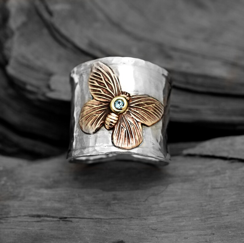 K&ouml;nnte beinhalten: Ein silberner Ring mit geh&auml;mmerter Textur zeigt ein detailliertes Schmetterlingsdesign mit einem blauen Edelstein in der Mitte. Der Schmetterling ist aus Kupfer und hat einen goldenen Akzent.