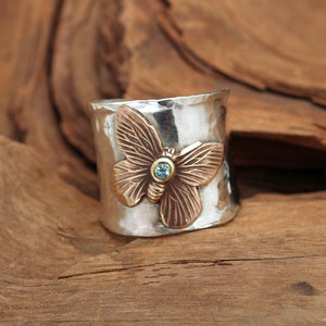 K&ouml;nnte beinhalten: Ein silberner Ring mit geh&auml;mmerter Oberfl&auml;che zeigt einen bronzenen Schmetterling mit einem blauen Edelstein in der Mitte.