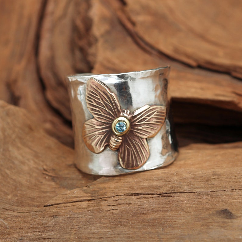 K&ouml;nnte beinhalten: Ein silberner Ring mit geh&auml;mmerter Oberfl&auml;che zeigt einen bronzenen Schmetterling mit einem blauen Edelstein in der Mitte.