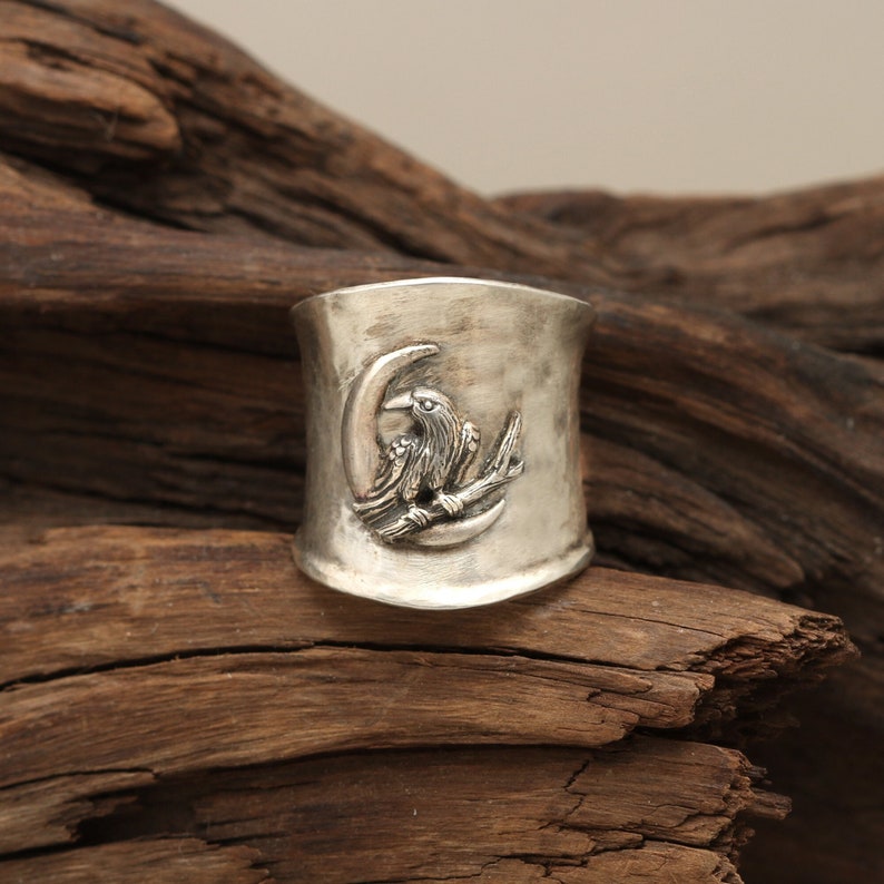 K&ouml;nnte beinhalten: Ein silberner Ring mit einem erhabenen Design eines Rabens, der auf einem Ast sitzt, mit einem Halbmond dahinter.