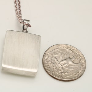 Op de afbeelding: Een zilveren ketting met een rechthoekige hanger. De hanger is ongeveer zo groot als een Amerikaanse kwart.