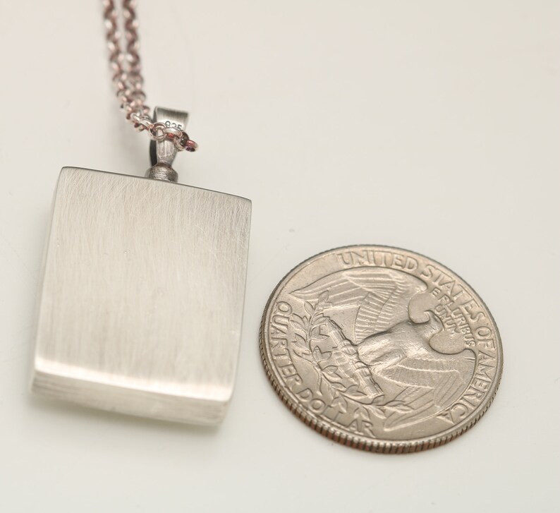 Op de afbeelding: Een zilveren ketting met een rechthoekige hanger. De hanger is ongeveer zo groot als een Amerikaanse kwart.