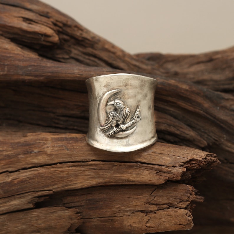 K&ouml;nnte beinhalten: Ein silberner Ring mit strukturierter Oberfl&auml;che zeigt ein erhabenes Design eines Rabens, der auf einem Halbmond sitzt.