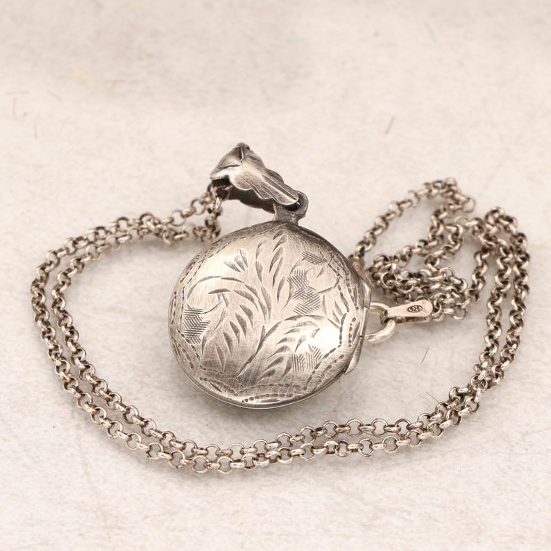 May include: A silver locket necklace with a delicate chain. The locket is round and features an engraved floral design.