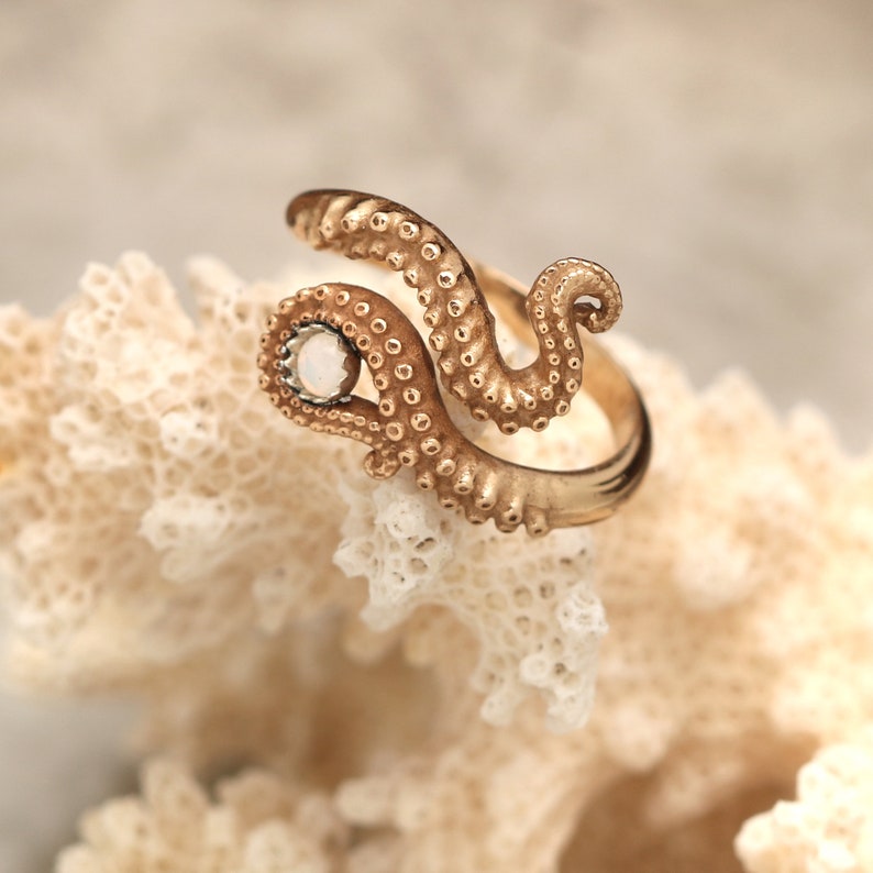 May include: A gold-toned ring shaped like an octopus tentacle with a textured surface featuring small suction cups. The ring has an open design and is accented with a single, small, round, white stone set at one end of the tentacle structure.