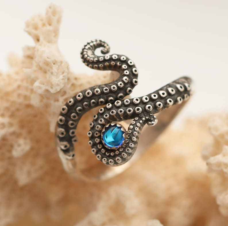 May include: A silver ring shaped like an octopus tentacle with a coiled design featuring a series of small circular suction cups along the band and a single round, bright blue gemstone set prominently near the center of the tentacle curve.