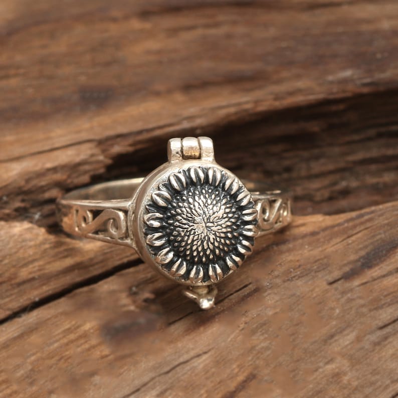 May include: A silver ring featuring a sunflower design. The ring has a hinged lid that opens to reveal a small compartment.