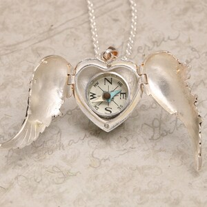 Pu&ograve; includere: Collana con ciondolo a forma di cuore in argento, un compasso all'interno e ali d'angelo su entrambi i lati. Il compasso ha un ago blu e un quadrante bianco con segni neri.