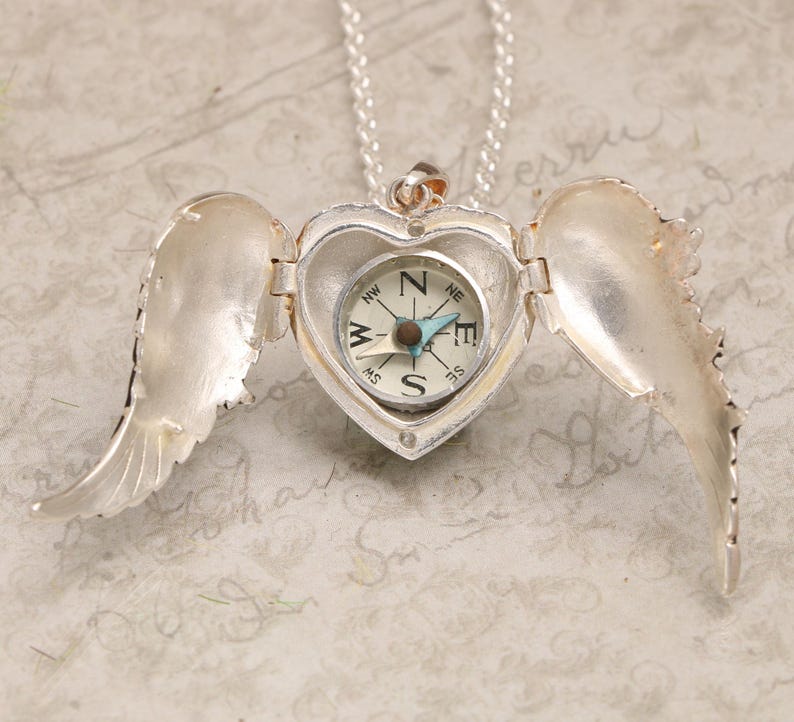 Pu&ograve; includere: Collana con ciondolo a forma di cuore in argento, un compasso all'interno e ali d'angelo su entrambi i lati. Il compasso ha un ago blu e un quadrante bianco con segni neri.