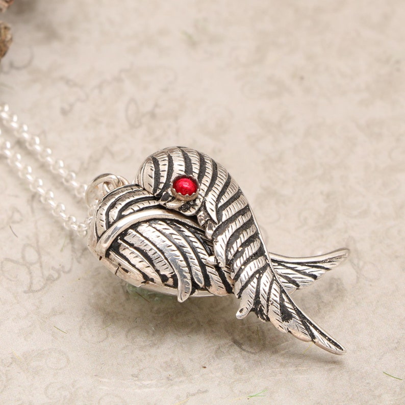 Pu&ograve; includere: Collana con ciondolo in argento a forma di cuore con un medaglione decorato con un paio di ali e una pietra preziosa rossa. Il medaglione &egrave; aperto e le ali sono spiegate.