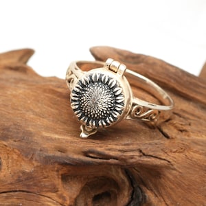 May include: A gold-toned ring with a hinged sunflower design. The ring is open and the sunflower is visible. The ring is on a piece of wood.