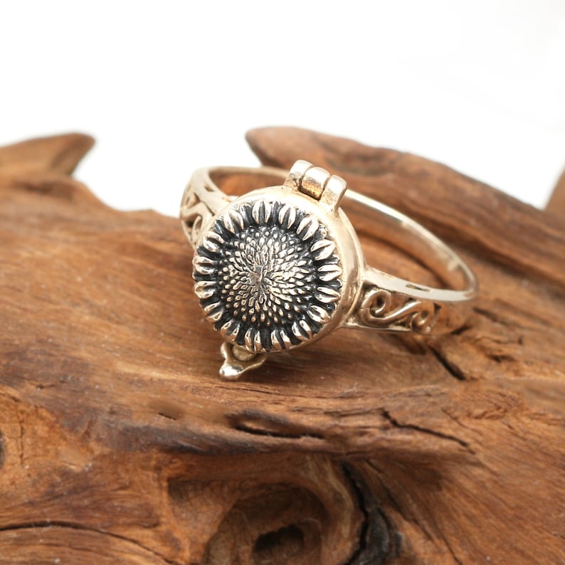 May include: A gold-toned ring with a hinged sunflower design. The ring is open and the sunflower is visible. The ring is on a piece of wood.