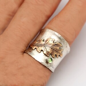 Puede incluir: Un anillo de plata y cobre con un acabado texturizado. El anillo presenta un dise&ntilde;o de hoja de roble en relieve con un acento de piedra preciosa verde.