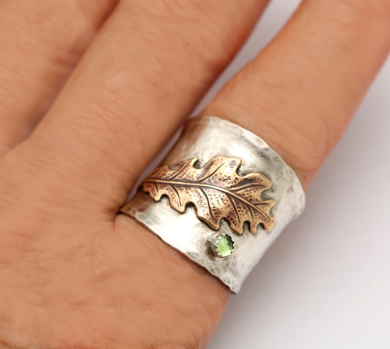 Puede incluir: Un anillo de plata y cobre con un acabado texturizado. El anillo presenta un dise&ntilde;o de hoja de roble en relieve con un acento de piedra preciosa verde.