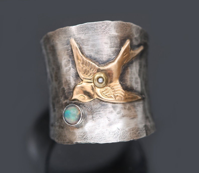 A sterling silver wide band ring on top that tapers smaller underneath. The width of the ring on top is the width of a nickel. The ring is accented with a gold filled soaring song bird with a tiny pearl in the center and a real opal below the bird.