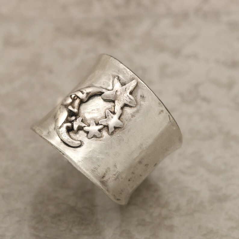 May include: A silver ring featuring a crescent moon and stars design. The ring is crafted from hammered metal and has a rustic appearance.