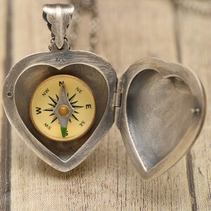 May include: A silver heart-shaped locket with a compass inside. The locket is open and the compass is visible. The compass has a gold face with black markings and a green needle.
