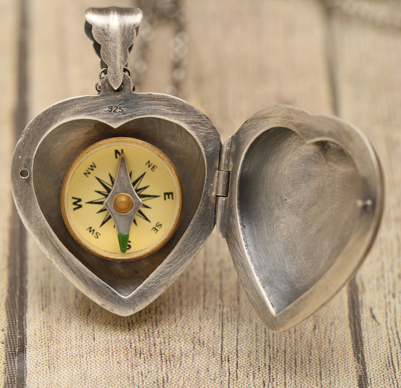 May include: A silver heart-shaped locket with a compass inside. The locket is open and the compass is visible. The compass has a gold face with black markings and a green needle.