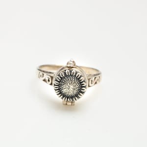 May include: A silver ring featuring a sunflower design. The ring has a hinged lid that opens to reveal a small compartment.