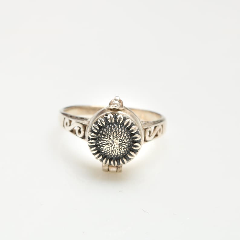 May include: A silver ring featuring a sunflower design. The ring has a hinged lid that opens to reveal a small compartment.