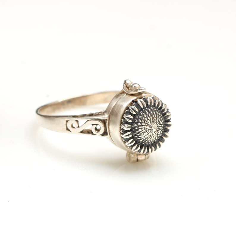 May include: A silver ring featuring a sunflower design. The ring is a hinged locket that opens to reveal a small compartment.